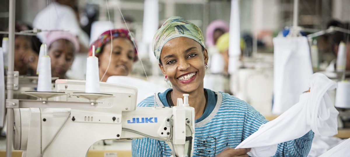 Ethiopische vrouw zit achter een naaimachine en kijkt lachend in de camera. Zij heeft een blauw gestreept shirt aan en heeft een stuk witte stof in haar handen.